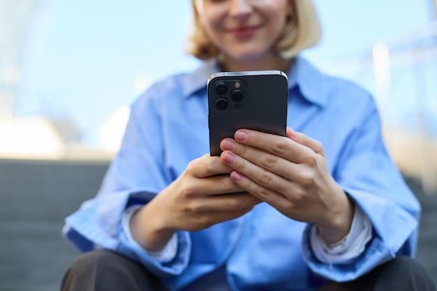 Close up portrait of young woman holding smartphone in both hands sitting on street with mobile