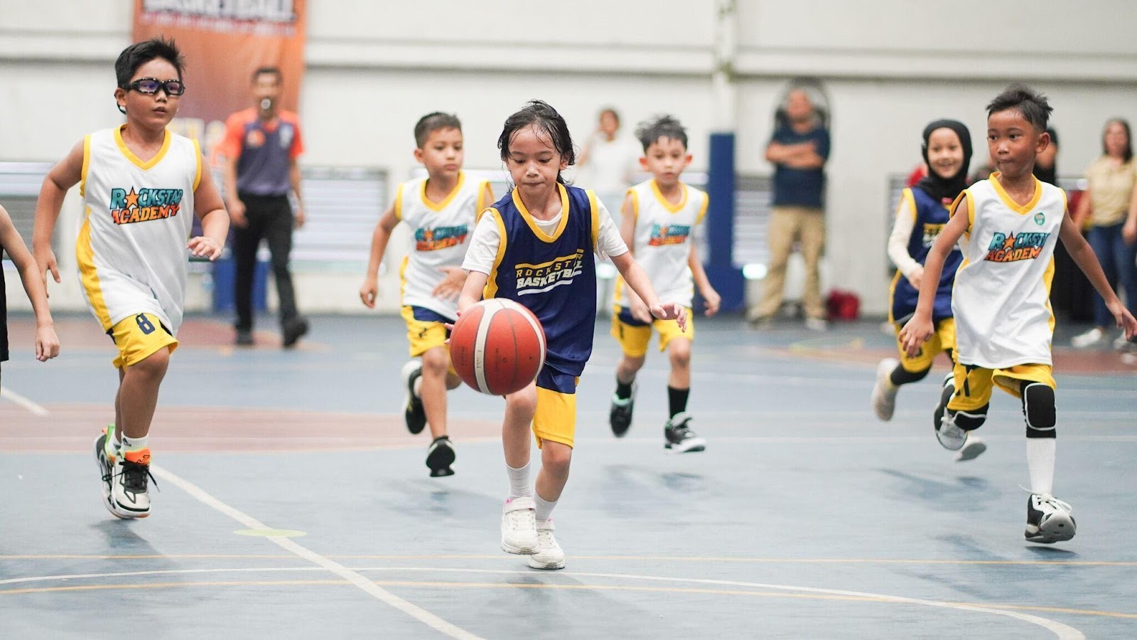 Anak perempuan dengan jersey biru men-dribble bola basket sambil dikelilingi oleh tim lawan