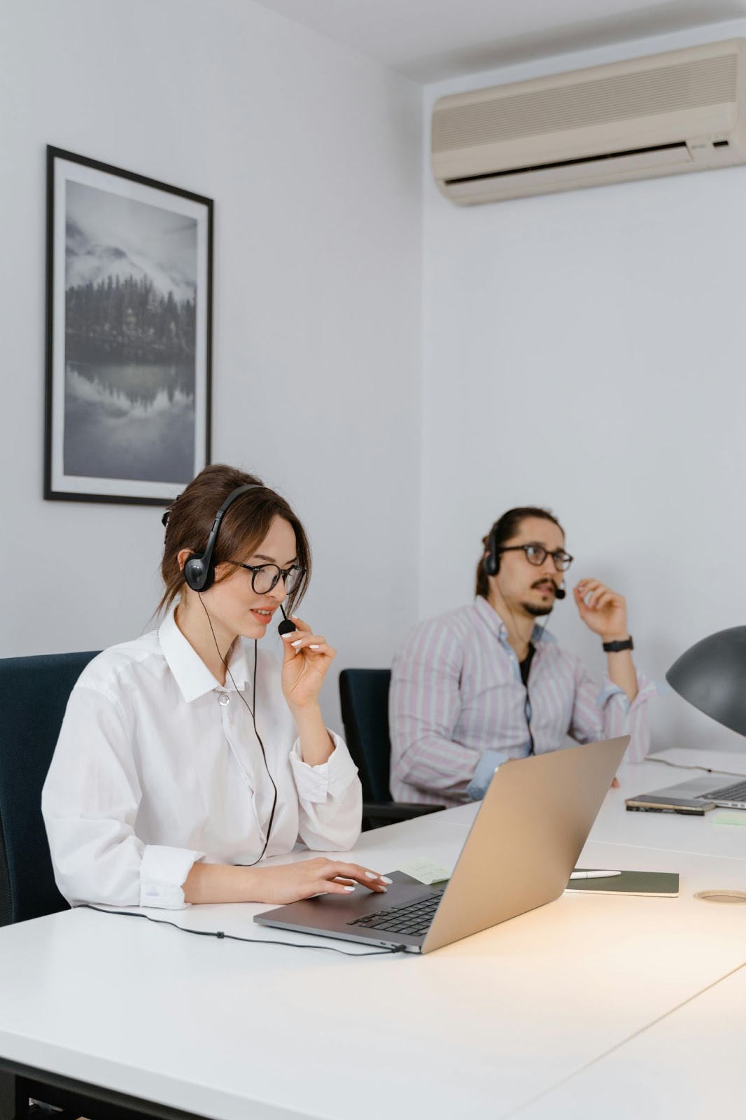 A man and a woman wearing headsets work on their laptops in an office, with the woman speaking into her headset.