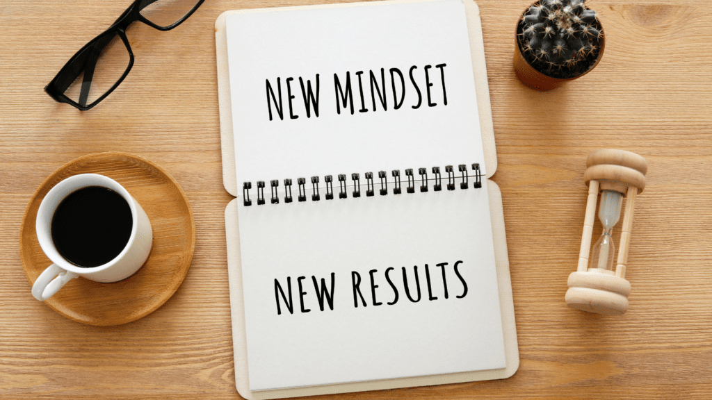 An open notebook on a wooden desk with the words New Mindset, New Results written inside, next to a cup of coffee and an hourglass.