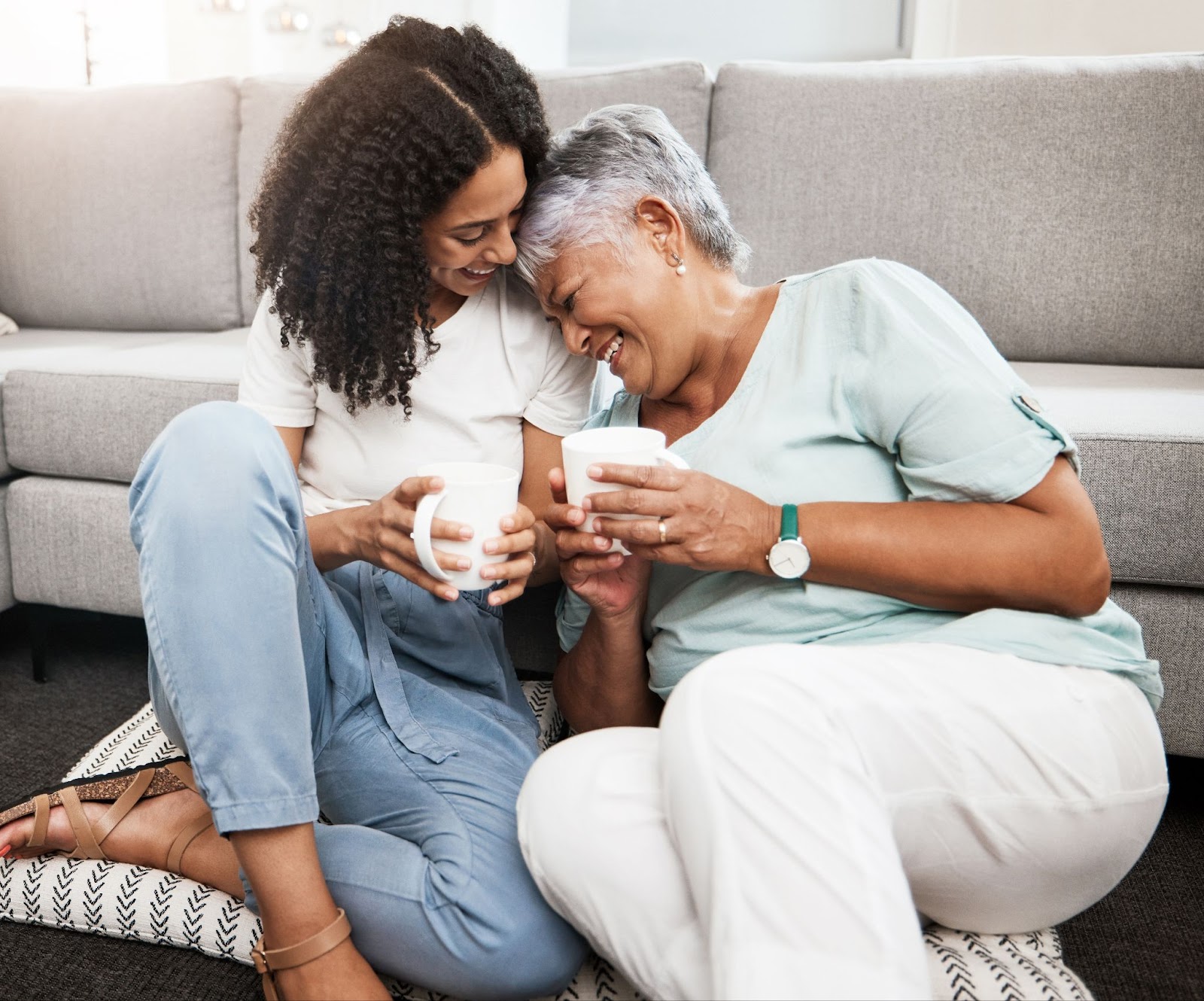 A senior woman and her daughter sitting in front of the couch while holding coffee and laughing during a visit.