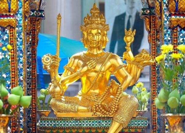 A gold statue of a person with Erawan Shrine in the backgroundDescription automatically generated