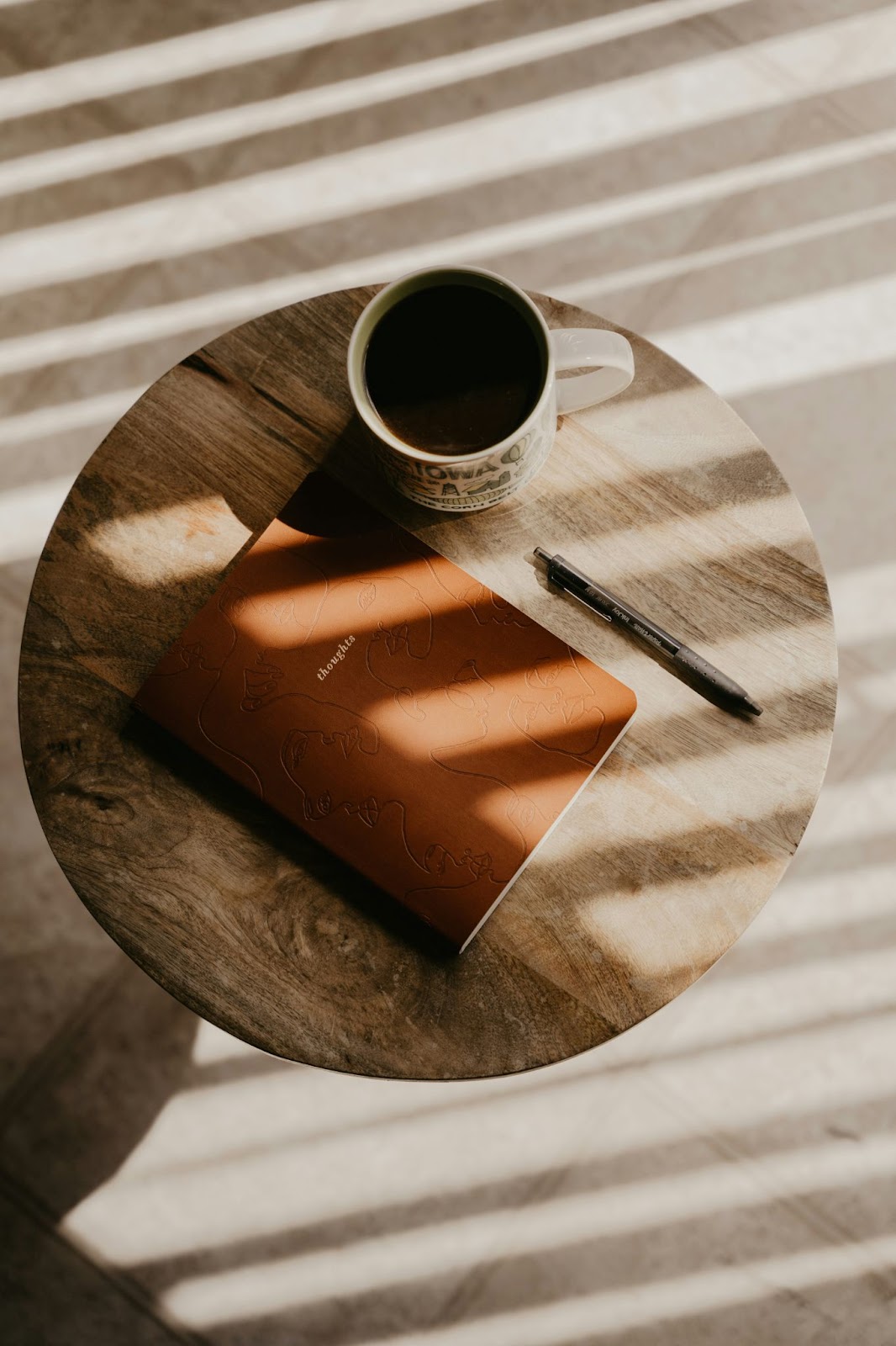 A round wooden side table with a cup of coffee, a closed brown journal, and a black pen resting on it. Sunlight filters through blinds, casting striped shadows across the scene.