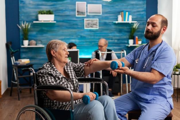 Male nurse helping senior retired disabled woman in wheelchair to rehabilitate using dumbbels