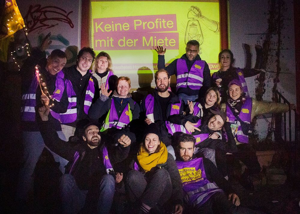 Organizers with Deutsche Wohnen & Co. Enteignen wearing purple vests from doorknocking stand pose under a sign that says "Keine Profite mit der Miete" (No Profit With the Rent)