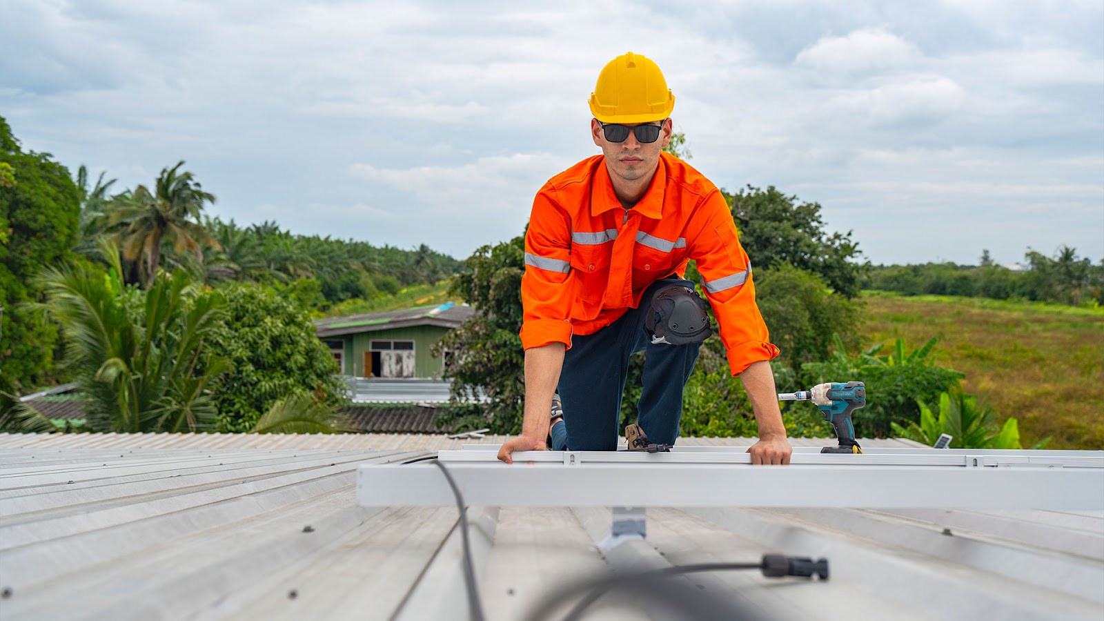 Technician in safety uniform and yellow helmet kneeling on a rooftop, inspecting and securing metal panels with a drill