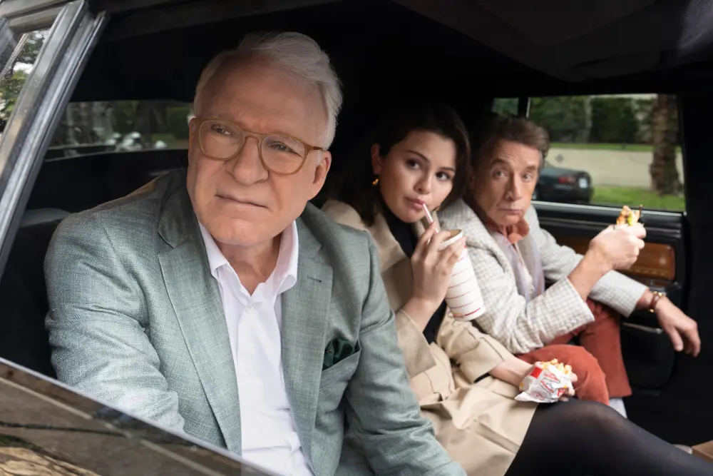 Personajes de 'Only Murders in the Building' interpretados por Steve Martin, Selena Gomez y Martin Short en un coche, tomando bebidas y snacks, escena cómica mientras discuten un caso.