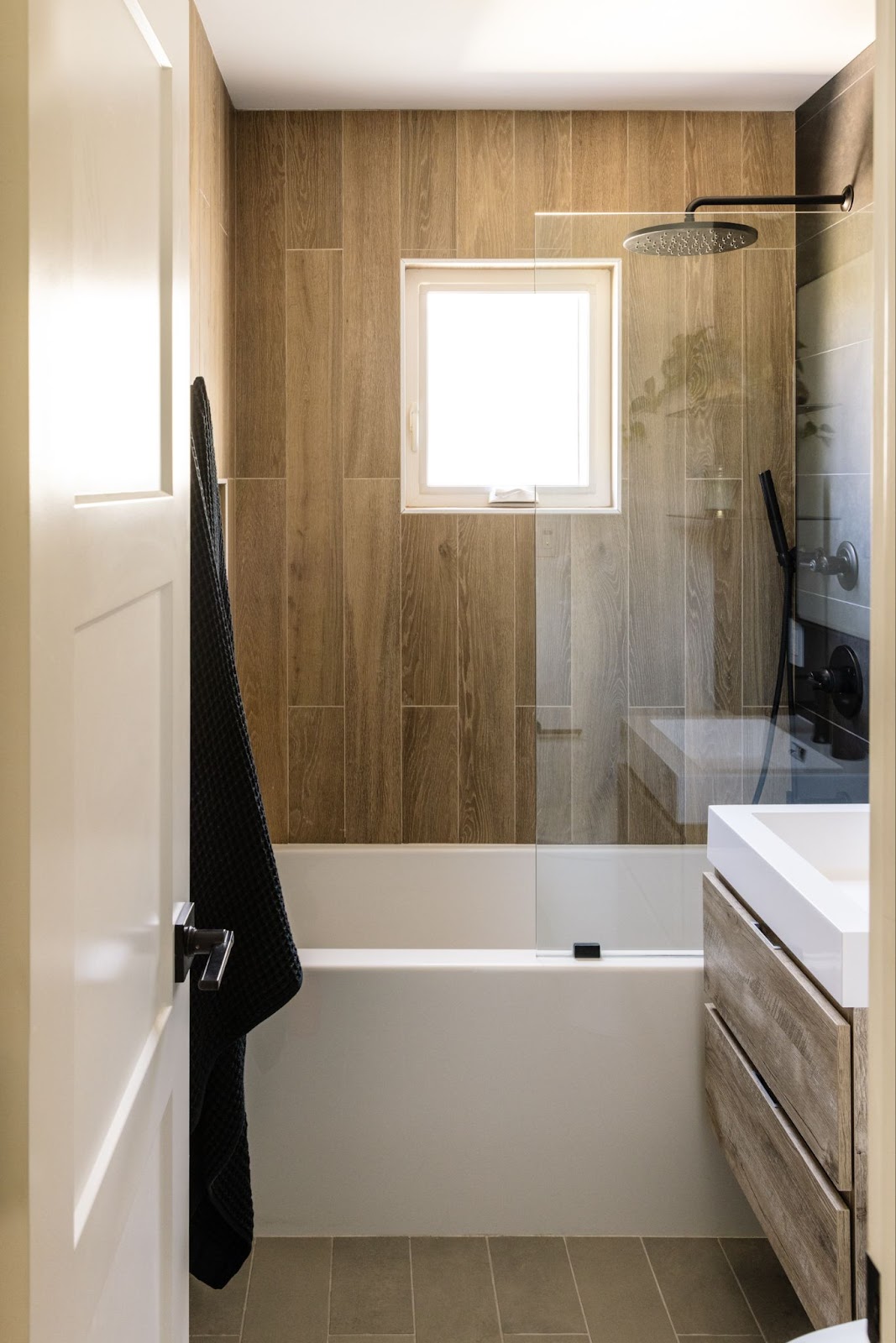 Small full-bath with tub-shower combo, wood grain tile wall panels, large format charcoal tile, and a wood vanity