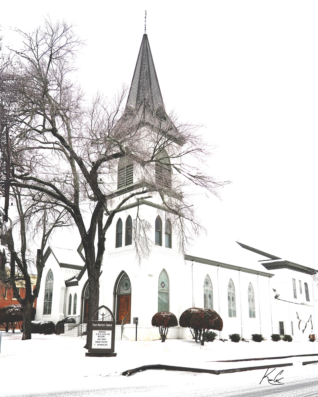 A beautiful white church with pointed arches and a tall steeple, surrounded by snow-covered grounds and trees. Captured by @thevalcoxgallery.