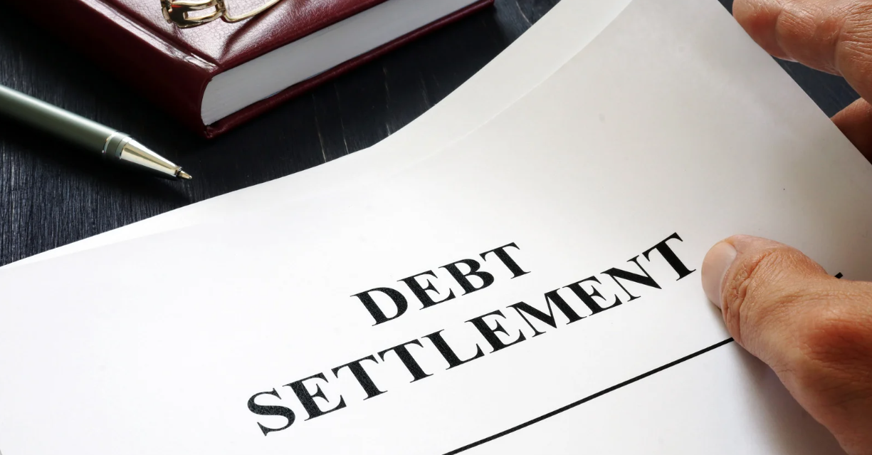(A close-up of a document titled “Debt Settlement” on a dark desk, with a hand holding the paper, a pen, and a notebook nearby.)