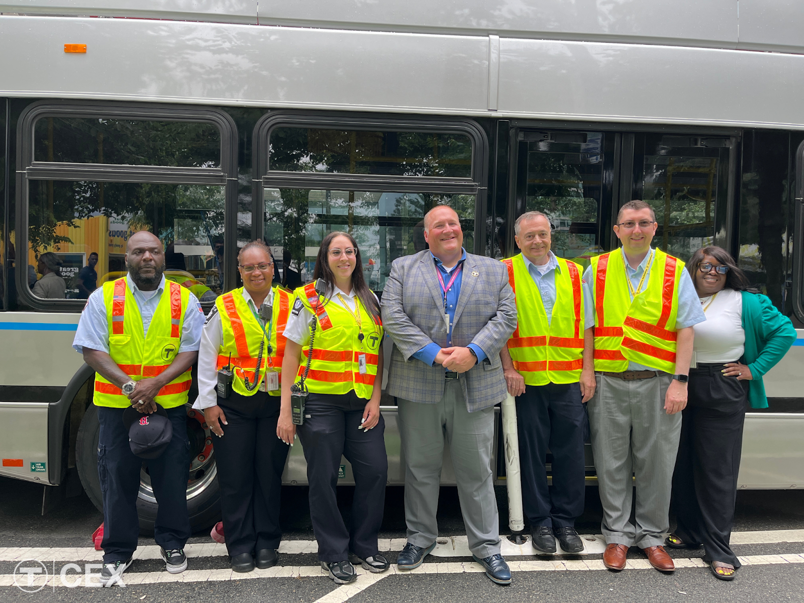 Seven individuals pose smiling in front of a bus. Several of the employees wear high visibility vests.