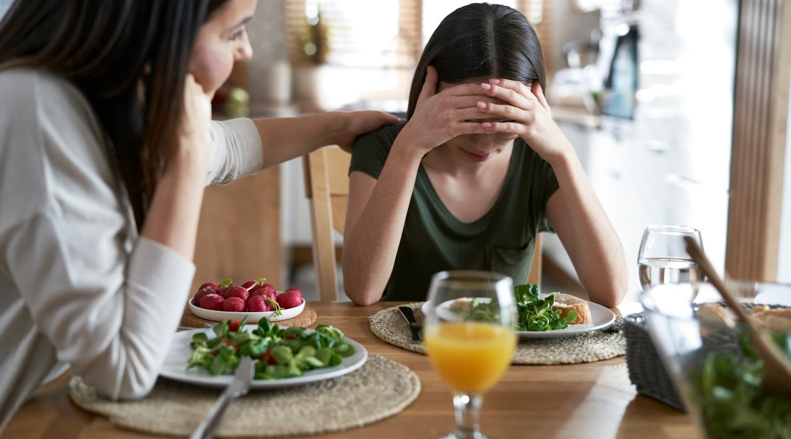 Woman comforting a distressed girl at the dining table, both seated in front of plates with salad and juice.