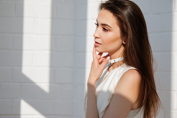 Young brunette female pictured from the chest up. She has long hair that looks healthy and in good condition, because she follows a good natural hair care routine. She is wearng a white sleevless top and is holdng her hand to her mouth. She is stood to the right of the photo against a white brick wall background with shadows.