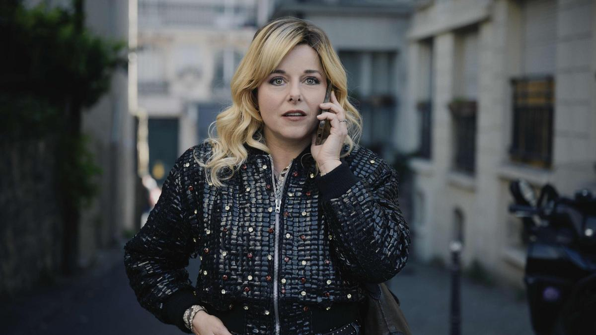 Mujer rubia con chaqueta negra de estilo acolchado, hablando por teléfono en una calle de París. Su expresión es de preocupación mientras sostiene el teléfono con una mano y camina por una calle estrecha, rodeada de edificios parisinos.