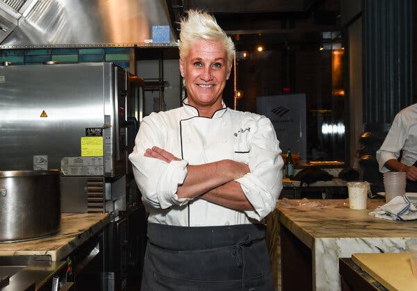 A woman with short, blond hair wearing a chef’s coat stands with her arms crossed in a restaurant kitchen.