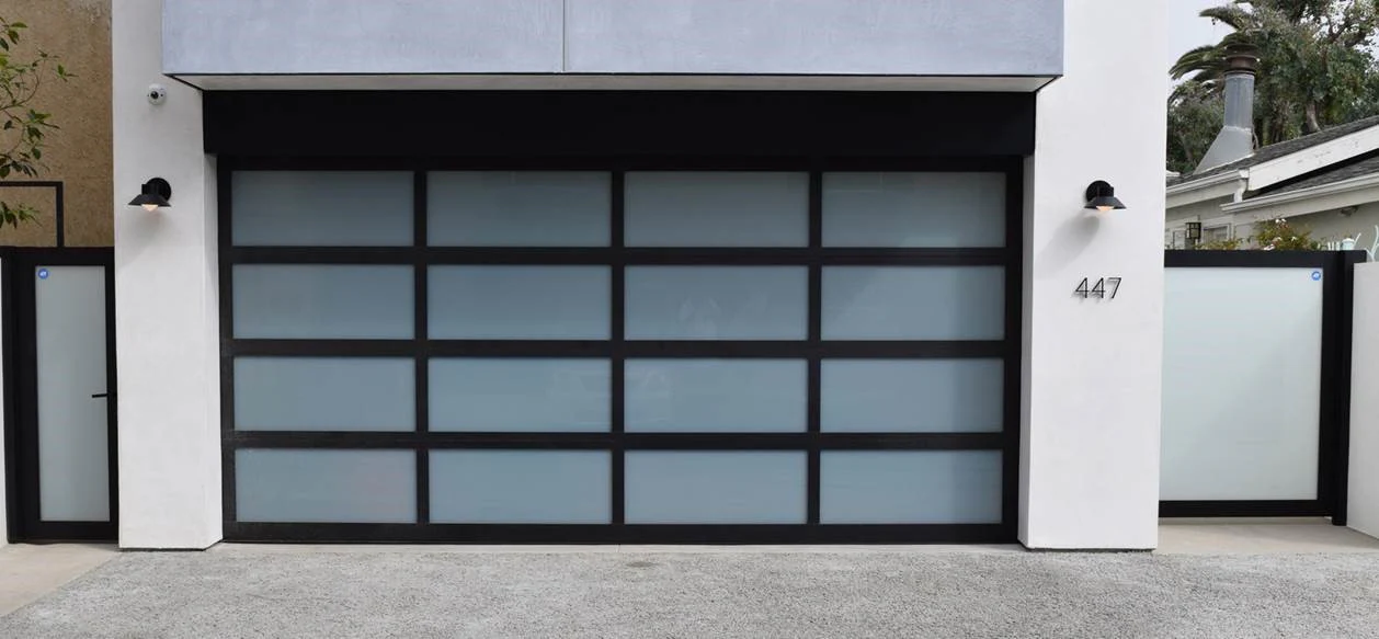 a sleek aluminum garage door with frosted glass panels arranged in a grid design, framed by black aluminum. The contemporary aesthetic complements the minimalist architecture of the surrounding home, featuring clean white walls and black accents