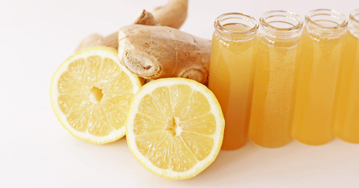 four mini glass bottles full of ginger shots with a lemon sliced in half and ginger root next to it