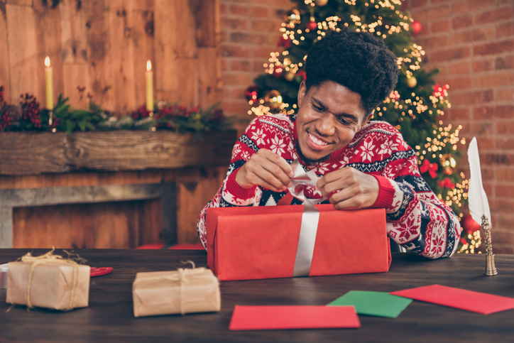 A man in a festive sweater unwrapping a present in front of a Christmas tree.