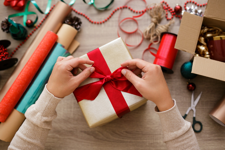 Two hands tie an elegant red bow onto a present, with holiday decorations scattered around. 