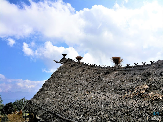 A traditional thatched roof with decorative elements, reminiscent of Nagaland architecture, stands under a partly cloudy blue sky.