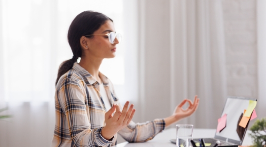 A woman wearing glasses and a plaid shirt, meditating in a seated position at her desk, hands resting on her knees, with a laptop and sticky notes in the background, in a bright and calm environment.