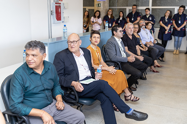 A imagem mostra um grupo de pessoas sentadas em cadeiras em um ambiente interno. Na fileira da frente, estão pessoas de aparência mais formal, vestidas com roupas sociais e elegantes. Um homem de terno escuro e óculos segura uma garrafa de água e está conversando ou atento a algo fora da imagem. Ao seu lado, uma mulher usando um vestido laranja e sandálias observa a mesma direção.  Ao fundo, há mais pessoas, algumas de pé, vestindo uniformes de trabalho azul marinho, possivelmente funcionárias do local. A atmosfera parece formal e relacionada a um evento ou encontro profissional, como uma inauguração, reunião ou visita técnica. O local aparenta ser limpo, organizado e iluminado, com um piso claro e paredes brancas.