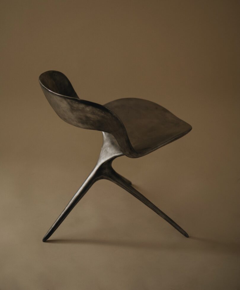 A modern black sculptural chair with a curved seat and backrest, supported by two angled legs, set against a plain brown background