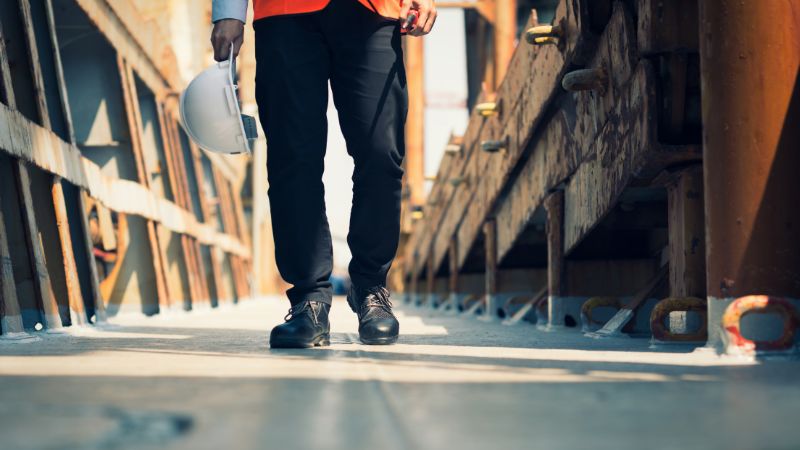 Person in black pants and work boots walking at port, holding a white hard hat.