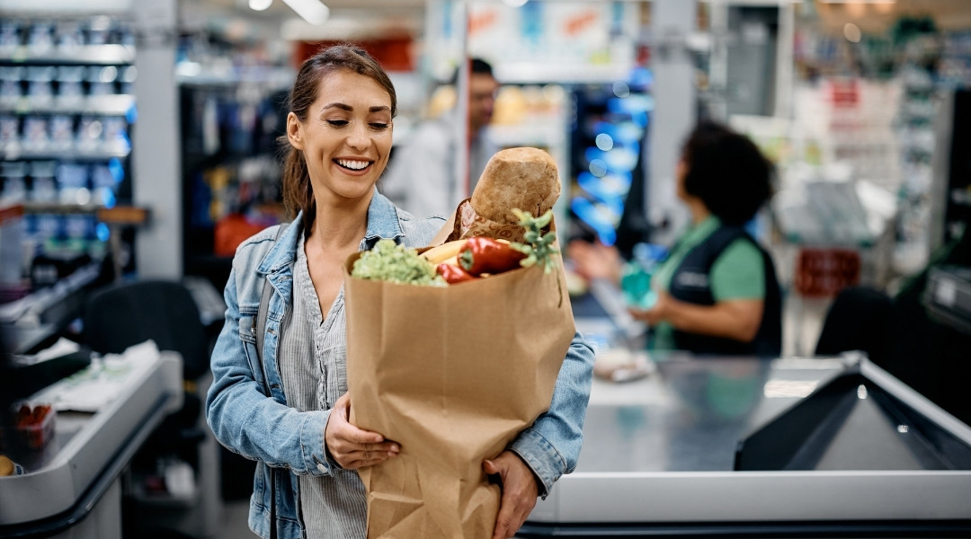 Une femme souriante tenant un sac en papier rempli de produits frais à la caisse d'une épicerie à Shawinigan.