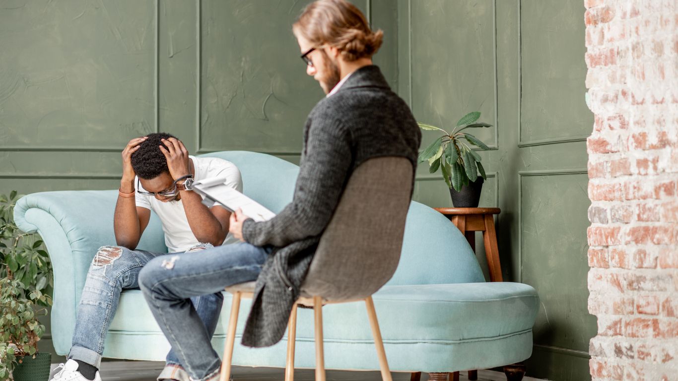 A therapy session where a young man appears stressed, sitting across from a therapist in a modern, comfortable setting