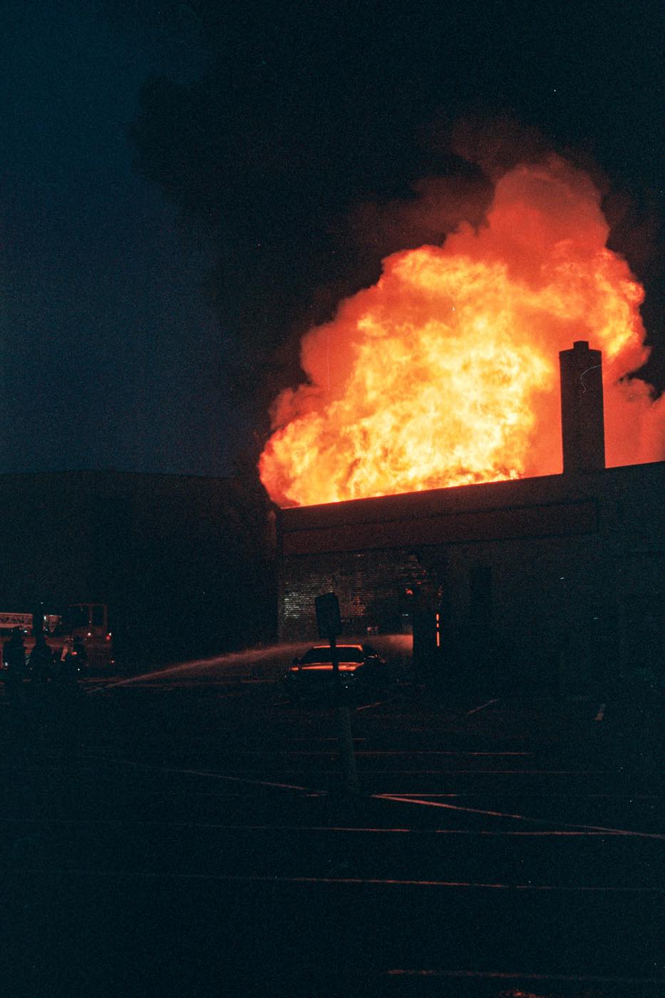 Image: Photo of Minneapolis uprising, 2020. The photo is mostly in darkness, with a fire ablaze in the top right corner, casting the building it is engulfing in some light. Photo sourced from CrimeThinc.com.