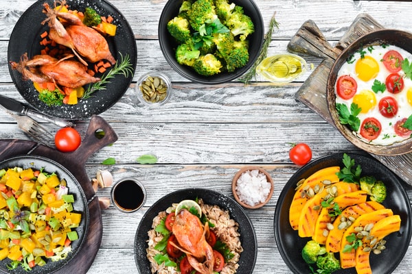 A meal spread featuring roasted quail, fresh salads, sautéed vegetables, eggs, and grains, with some dishes prepared using an Instant Pot, all arranged on rustic wooden surfaces