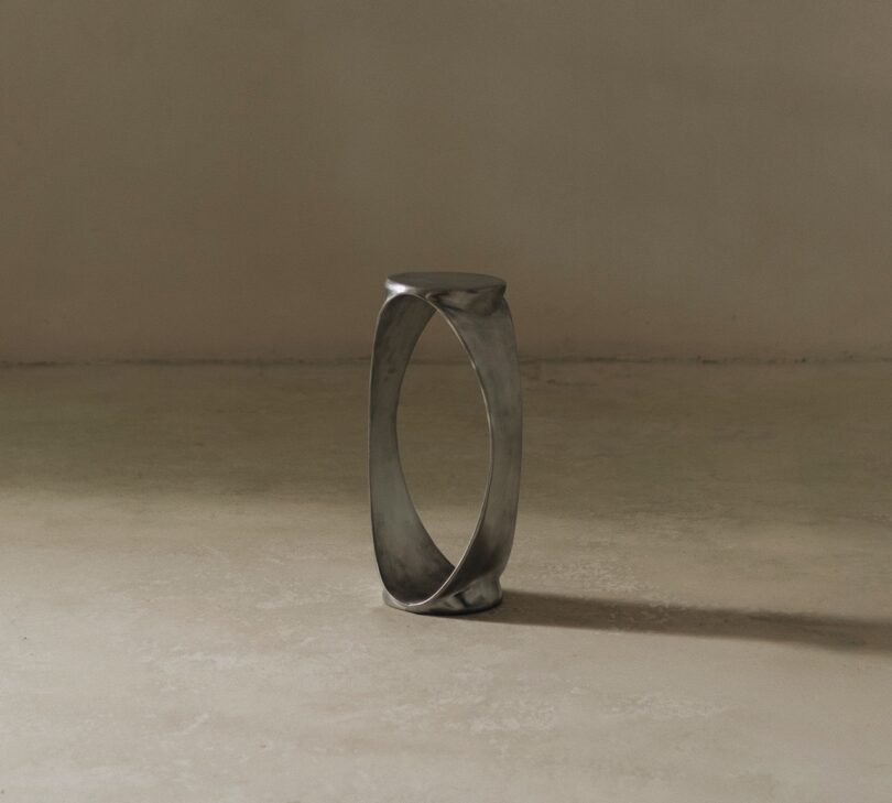A silver, oval-shaped metal table stands upright on a plain, light-colored floor against a neutral beige wall, casting a shadow.