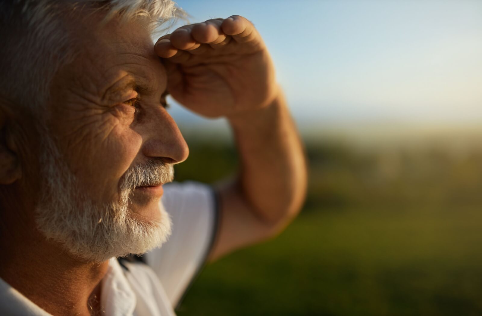 Side-view of a man with a white beard looking into the sun at golden hour and holding his left hand up to shade his eyes