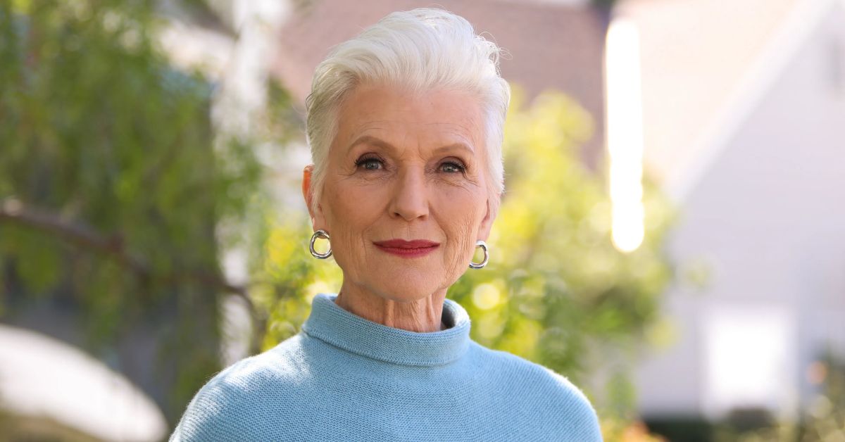 An older woman with a short spiky pixie cut, wearing a blue sweater, smiles warmly at the camera.