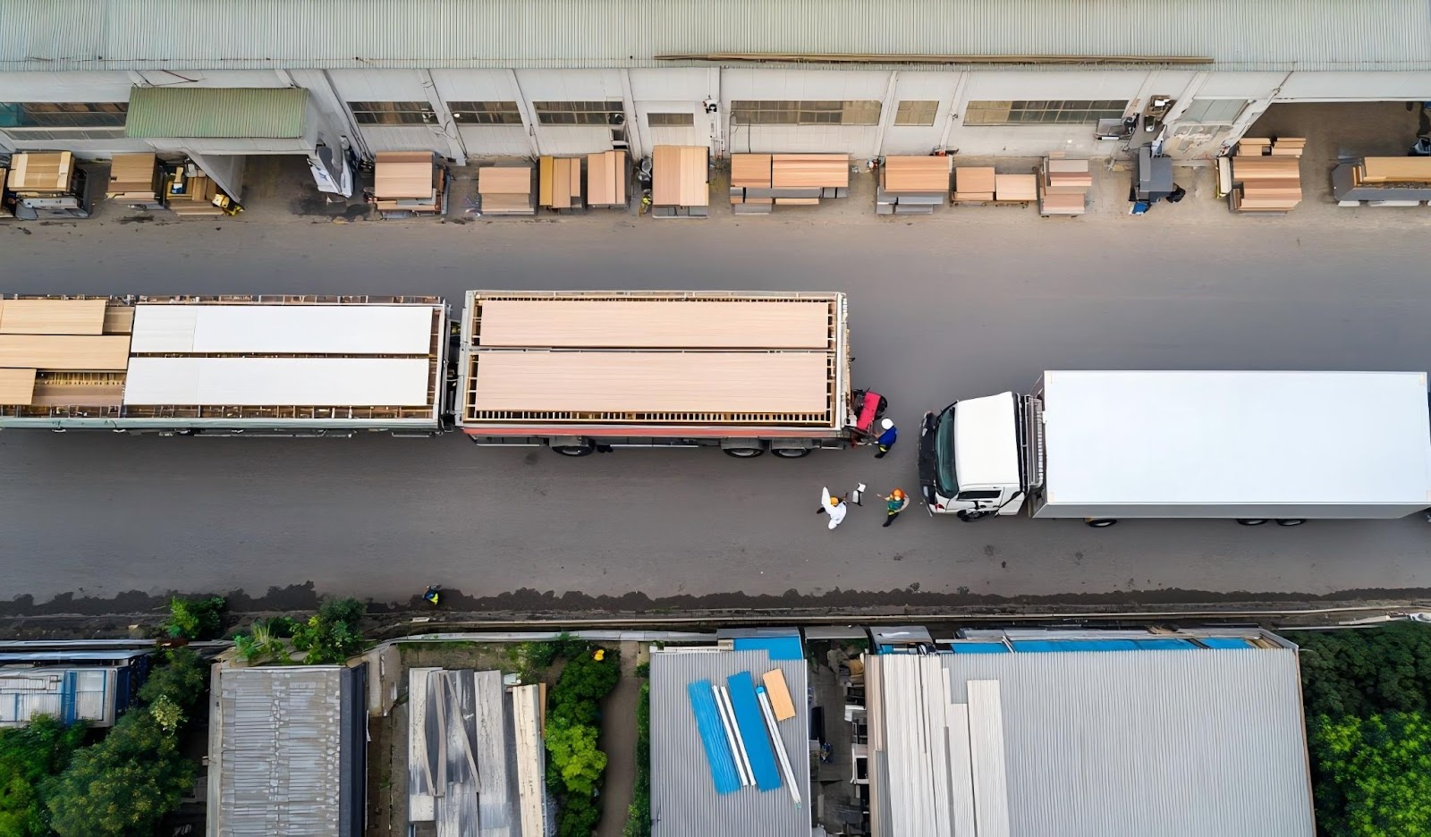 Aerial view of cabinet manufacturing logistics with flatbed trucks loading wood panels outside a production facility in an industrial park.