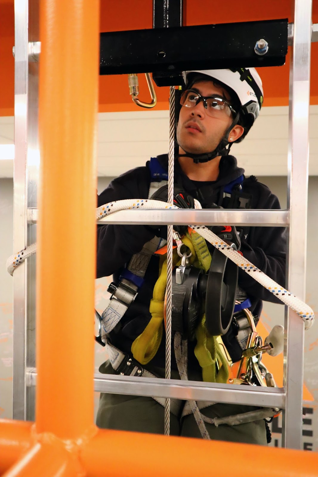 A close-up of an individual in safety gear, including a helmet, gloves, and harness, focused on operating a rope system during training. The orange railing frames the scene, highlighting a professional and secure environment.