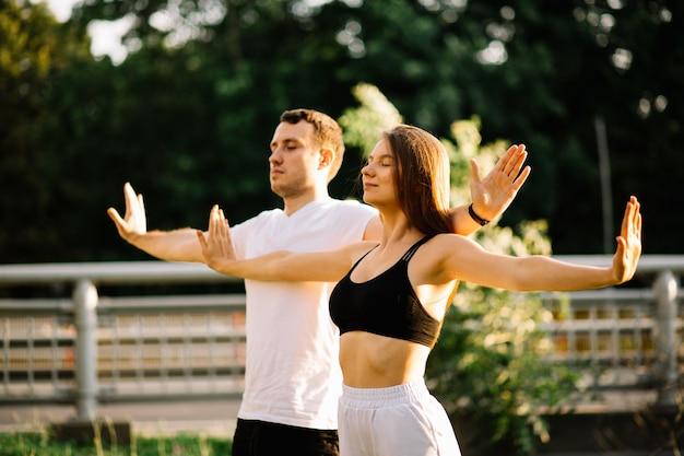 Young couple man and woman meditate together on sunset while standing, yoga on city lawn, summer evening, lifestyle, breathing exersices