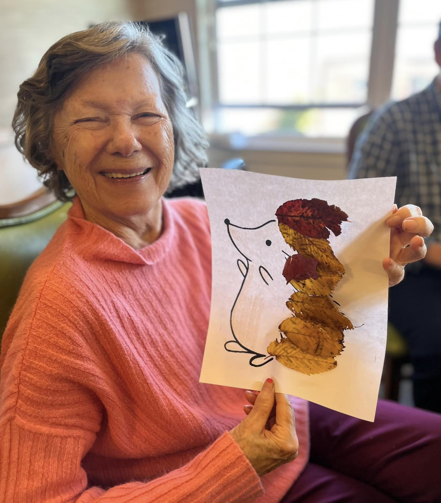 An assisted living resident holding up a piece of artwork of a squirrel with leaves pasted to its back