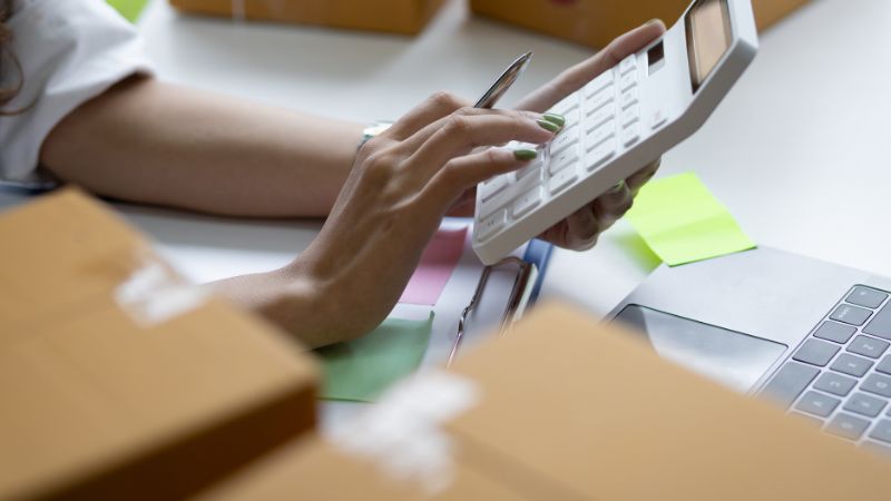 Person using a calculator at a desk with cardboard boxes and a laptop.