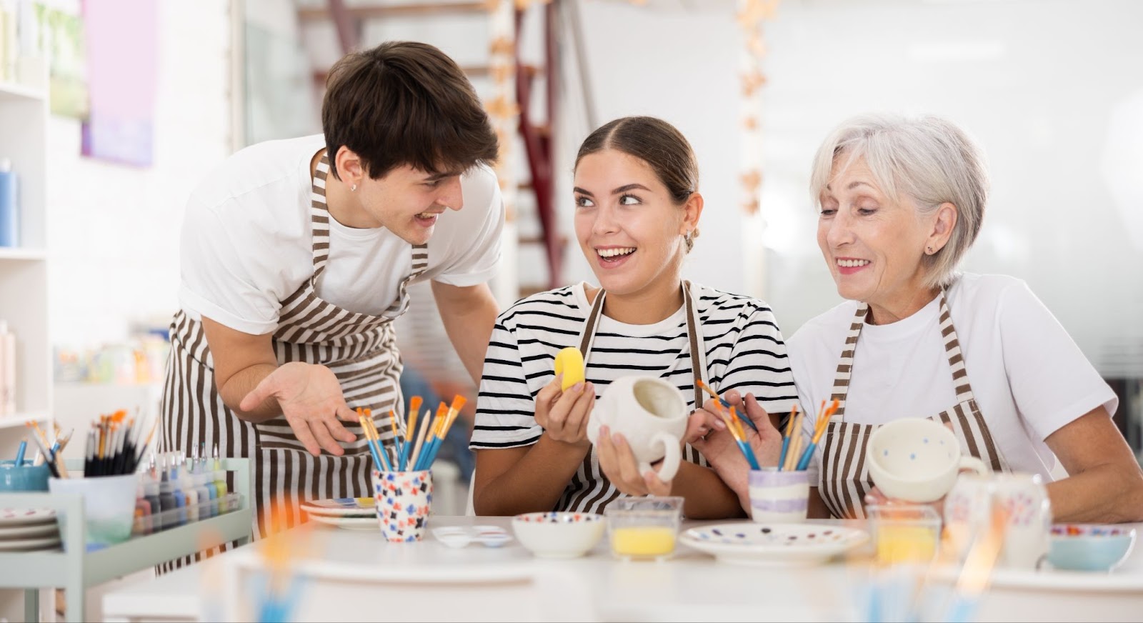 Two siblings sitting with their elderly parent painting mugs together in a brightly lit room.