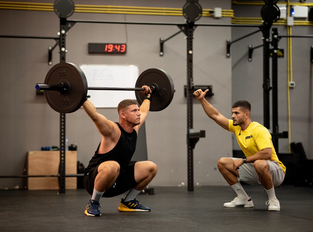 Free Athlete performing weightlifting exercise under trainer's guidance in a gym setting. Stock Photo