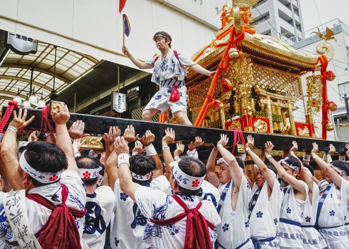 Many festival participants carrying a mikoshi portable shrine with one person standing on top to direct