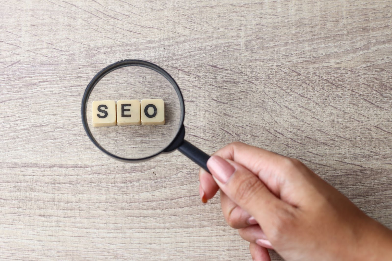 Woman with painted nails holding a magnifying glass over the word “SEO” written on tiles. 