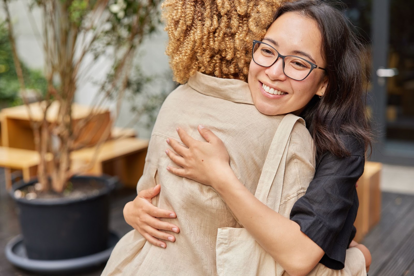 Two women hugging next to a table and a potted plant.