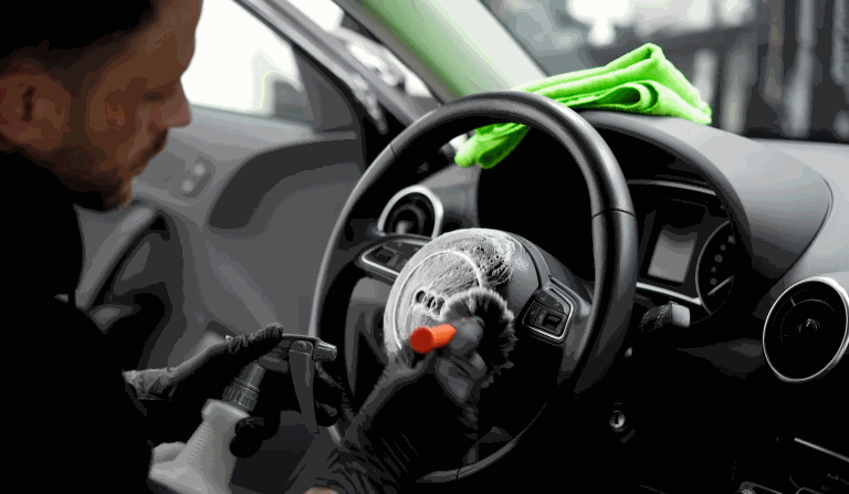 A person using a detailing brush with foam cleaner to meticulously clean a car's steering wheel. A green microfiber cloth is draped over the dashboard, indicating a car interior cleaning process.