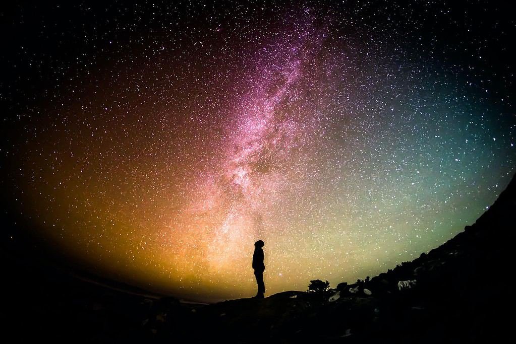 The 8th Chakra: A silhouette of a person standing on a rocky ground against a magnificent night sky filled with stars. The Milky Way is prominently visible, displaying a spectrum of colors from pink and purple to blue and green, creating an awe-inspiring backdrop.