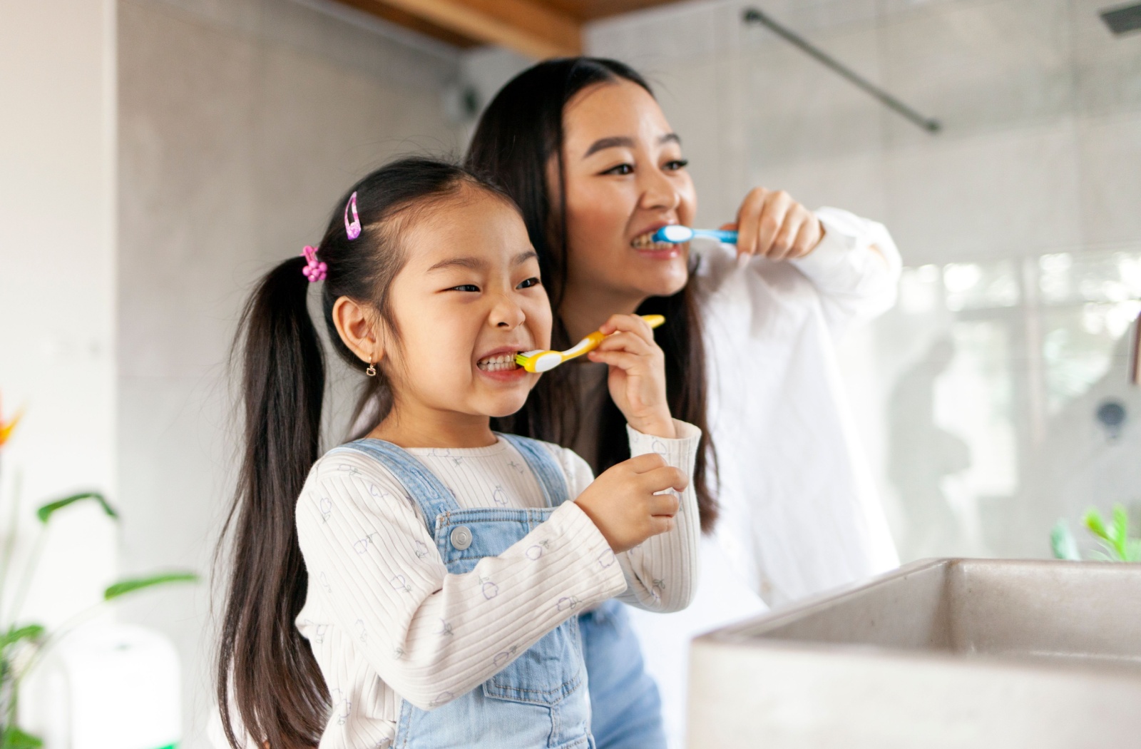 A child and an adult brushing their teeth together in a bathroom, smiling and practicing good oral hygiene.