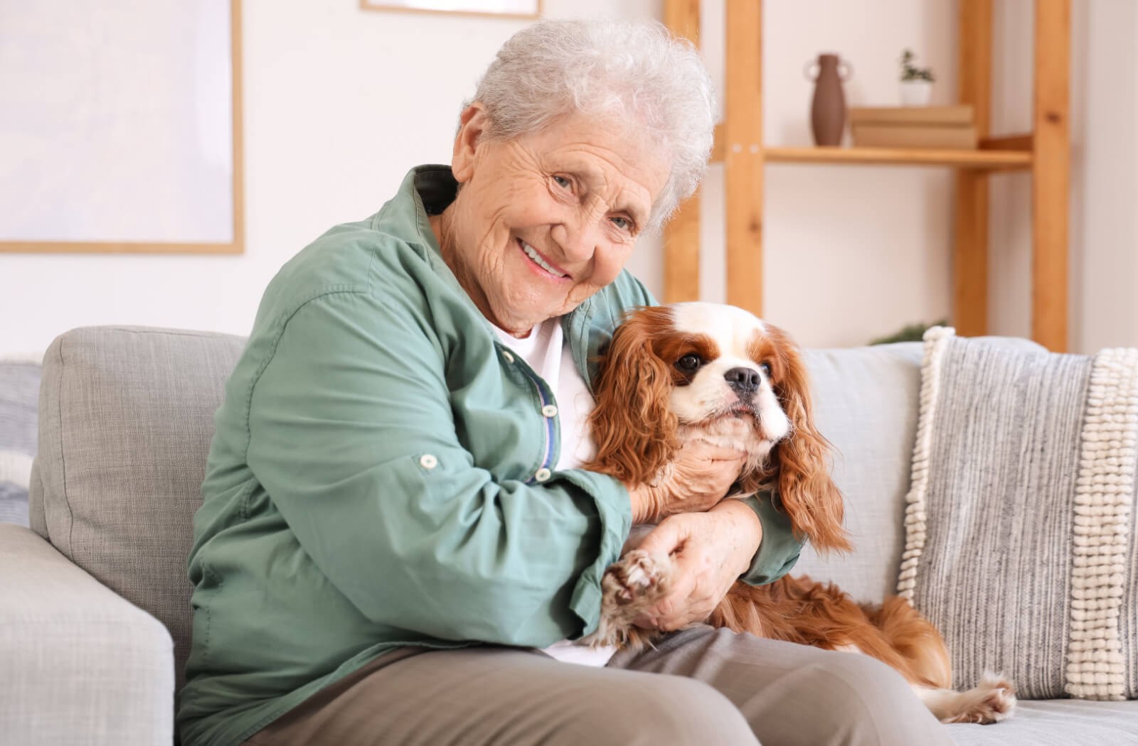 An older adult smiling while hugging their Cavalier King Charles Spaniel on the couch in senior living.