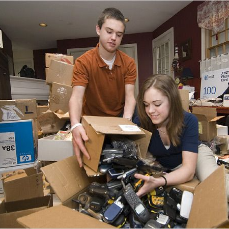 Two young volunteers sorting a large box of donated old cell phones for recycling